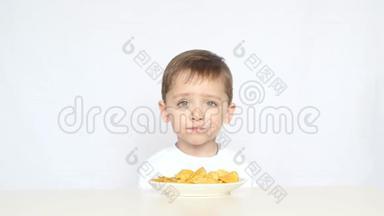 一个可爱的小男孩坐在桌子前的一盘薯片在白色的背景上。 孩子吃薯片